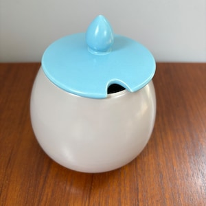 May include: A white ceramic sugar bowl with a light blue lid and a small spout. The lid has a pointed top.