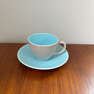 May include: A grey teacup with a light blue interior sits on a matching saucer. The cup has a curved handle and a smooth, glossy finish. The saucer and cup are on a wooden surface.
