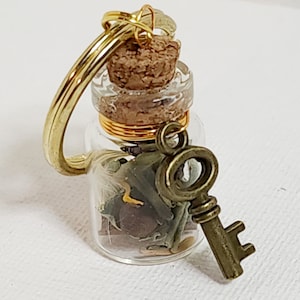 Könnte beinhalten: Eine kleine Glasflasche mit einem Korkverschluss und einem goldenen Schlüsselanhänger. Die Flasche ist mit getrockneten Kräutern und anderen natürlichen Elementen gefüllt.