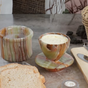 May include: A set of two green and brown marble butter dishes. The smaller dish is filled with butter and has a matching saucer. The larger dish is empty. There are slices of bread in the foreground.