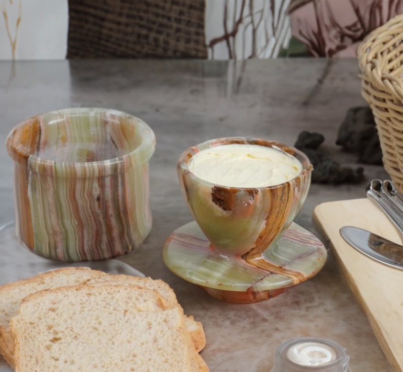 May include: A set of two green and brown marble butter dishes. The smaller dish is filled with butter and has a matching saucer. The larger dish is empty. There are slices of bread in the foreground.