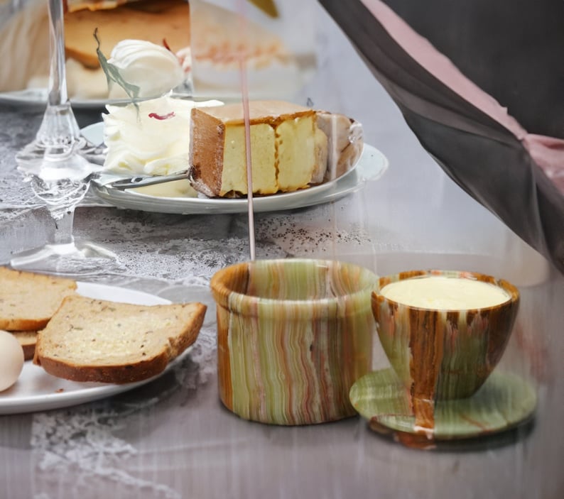 May include: Two green and brown striped ceramic butter dishes with lids. One dish is filled with a light yellow substance. The dishes are on a white surface with a piece of cake and bread.