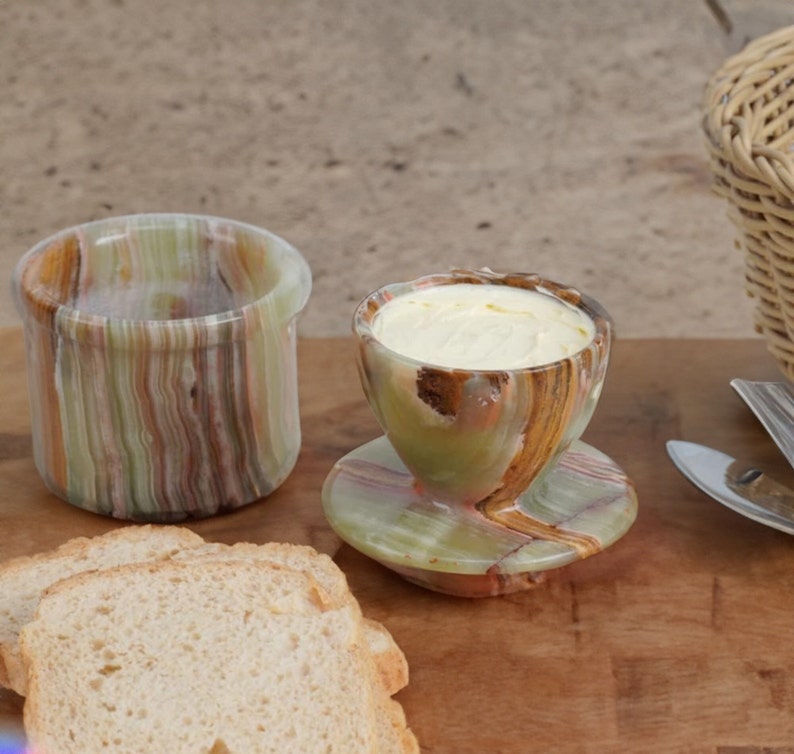 May include: A set of two green and brown marble butter dishes. The smaller dish has a lid and is filled with butter. The larger dish is empty and has a smooth, polished surface.