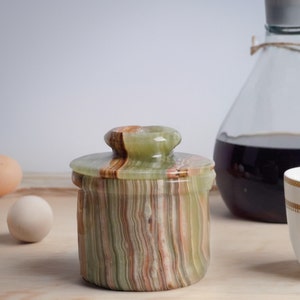 May include: A green, brown, and white striped onyx butter dish with a lid. The dish is sitting on a wooden surface.