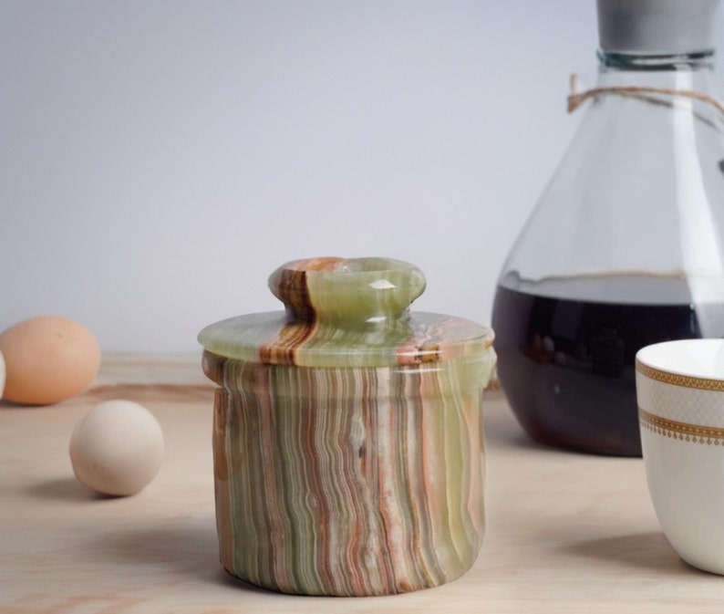 May include: A green, brown, and white striped onyx butter dish with a lid. The dish is sitting on a wooden surface.
