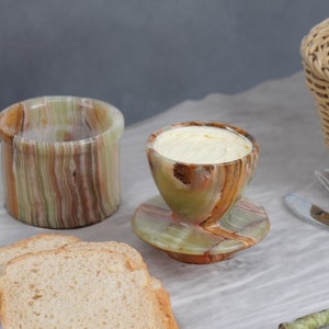 May include: A set of green and brown marble butter dishes. The butter dish has a lid and a small dish for serving butter. The dishes are on a white linen tablecloth with two slices of bread.