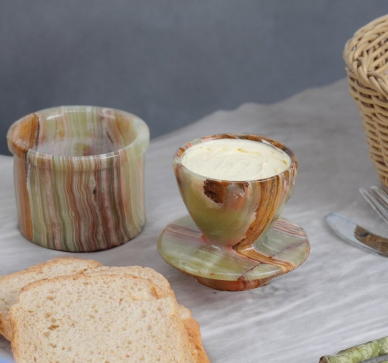 May include: A set of green and brown marble butter dishes. The butter dish has a lid and a small dish for serving butter. The dishes are on a white linen tablecloth with two slices of bread.