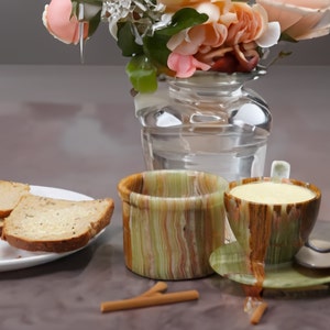 May include: Two striped ceramic mugs, one green and one brown, with a saucer. The green mug has a light yellow liquid inside. The brown mug is empty. The mugs are on a table with a vase of pink flowers in the background.