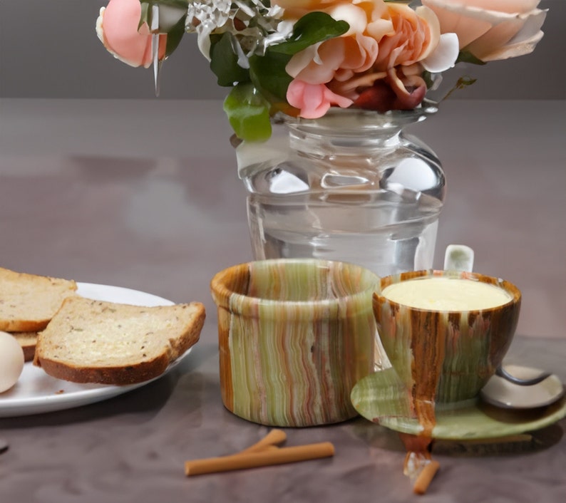 May include: Two striped ceramic mugs, one green and one brown, with a saucer. The green mug has a light yellow liquid inside. The brown mug is empty. The mugs are on a table with a vase of pink flowers in the background.
