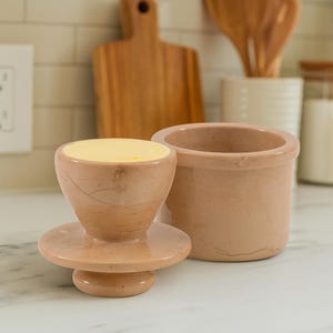 以下が含まれることがあります： 2 ピースのバター皿セット。バター皿は、明るい茶色の大理石のような素材で作られています。蓋は、平らなベースを持つ小さな逆円錐形です。ベースは円筒形の容器です。バター皿には黄色のバターが入っています。