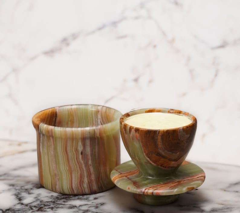 May include: A set of two green, brown, and white marble butter dishes. The smaller dish has a round shape and is filled with a pale yellow substance. The larger dish is rectangular and has a slightly wider opening.