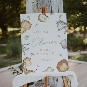 May include: A white sign with a watercolor illustration of animals, including a zebra, giraffe, lion, and tiger. The sign reads "Welcome to Olivia's Baby Shower 06.05.2023". The sign is on a wooden easel.