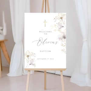Puede incluir: Un letrero blanco sobre un caballete de madera con el texto "Welcome to Olivia's Baptism" en escritura elegante. Una cruz dorada y detalles florales adornan la parte superior y los lados. La fecha "September 17, 2022" se muestra en la parte inferior.
