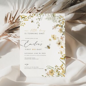K&ouml;nnte beinhalten: Eine wei&szlig;e Einladung mit einem floralen Rand in Gelb- und Gr&uuml;nt&ouml;nen. Der Text lautet "our little honey is turning one!" und "Please join us for Emilia's First Bee-Day!" Das Datum ist Samstag, der 18. Juni 2022.