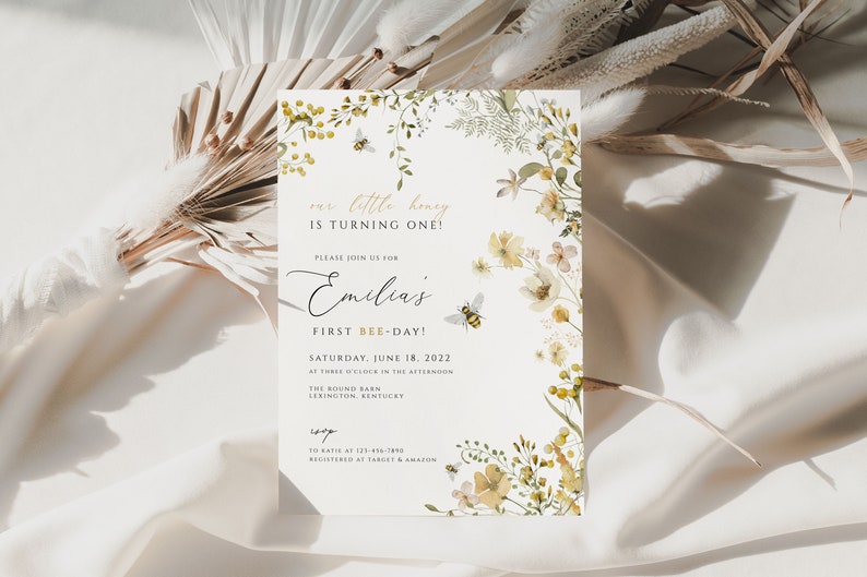 K&ouml;nnte beinhalten: Eine wei&szlig;e Einladung mit einem floralen Rand in Gelb- und Gr&uuml;nt&ouml;nen. Der Text lautet "our little honey is turning one!" und "Please join us for Emilia's First Bee-Day!" Das Datum ist Samstag, der 18. Juni 2022.