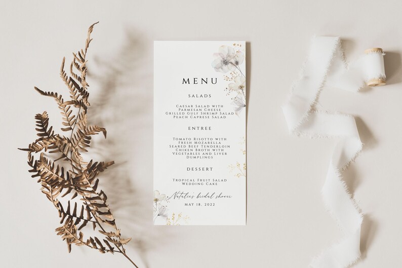 Puede incluir: Una tarjeta de men&uacute; blanca con un dise&ntilde;o floral y detalles dorados. El men&uacute; incluye ensaladas, plato principal, postre y el texto "Natasha's bridal shower May 18, 2022".