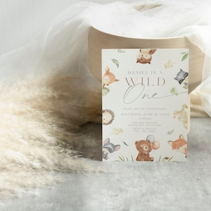 May include: A white invitation card with a woodland theme. The card features a brown bear, a fox, an owl, and a raccoon. The text on the card reads "Daniel is a Wild One" and includes details for a first birthday party.