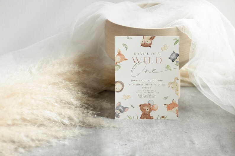 May include: A white invitation card with a woodland theme. The card features a brown bear, a fox, an owl, and a raccoon. The text on the card reads "Daniel is a Wild One" and includes details for a first birthday party.