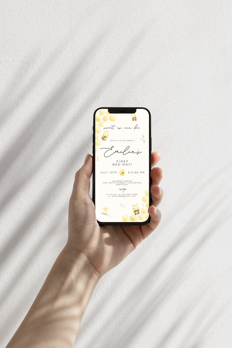 Op de afbeelding: Een smartphone die een uitnodiging voor een eerste verjaardagsfeestje met een bijen-thema voor Emilia laat zien. De uitnodiging heeft een honingraatpatroon, bijen en de tekst "sout ex cen bu" en "Vier met ons Emilias eerste bijendag!" De uitnodiging bevat ook de datum, tijd en locatie van het feest.