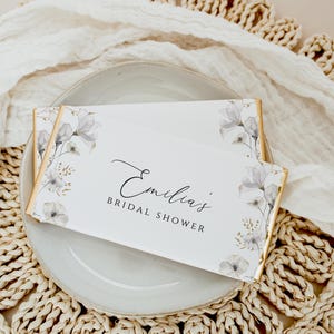 Puede incluir: Barra de chocolate blanco de favor con envoltorio de l&aacute;mina dorada y dise&ntilde;o floral. La barra est&aacute; etiquetada como "Ducha nupcial de Emilia".