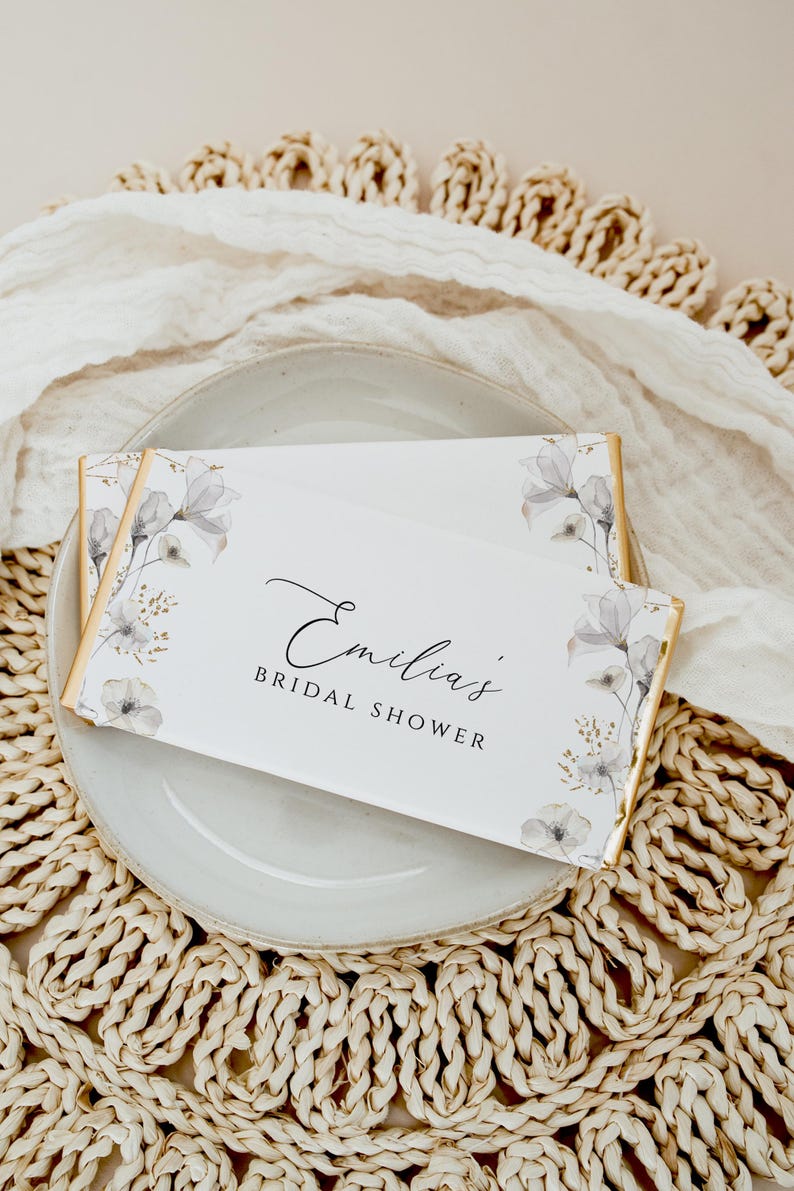 Puede incluir: Barra de chocolate blanco de favor con envoltorio de l&aacute;mina dorada y dise&ntilde;o floral. La barra est&aacute; etiquetada como "Ducha nupcial de Emilia".