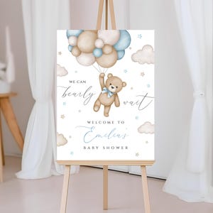 May include: A white welcome sign with a teddy bear holding balloons. The sign says "We can bearly wait" and "Welcome to Emilias Baby Shower".