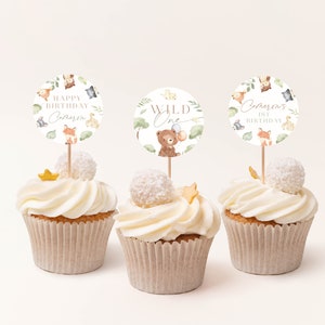 May include: Three cupcakes decorated with white frosting and topped with a round, white cake topper. The toppers feature a woodland animal theme with the text "Happy Birthday [Name] 1st Birthday", "Wild One", and "[Name] 1st Birthday".