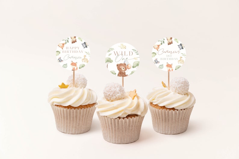 May include: Three cupcakes decorated with white frosting and topped with a round, white cake topper. The toppers feature a woodland animal theme with the text "Happy Birthday [Name] 1st Birthday", "Wild One", and "[Name] 1st Birthday".
