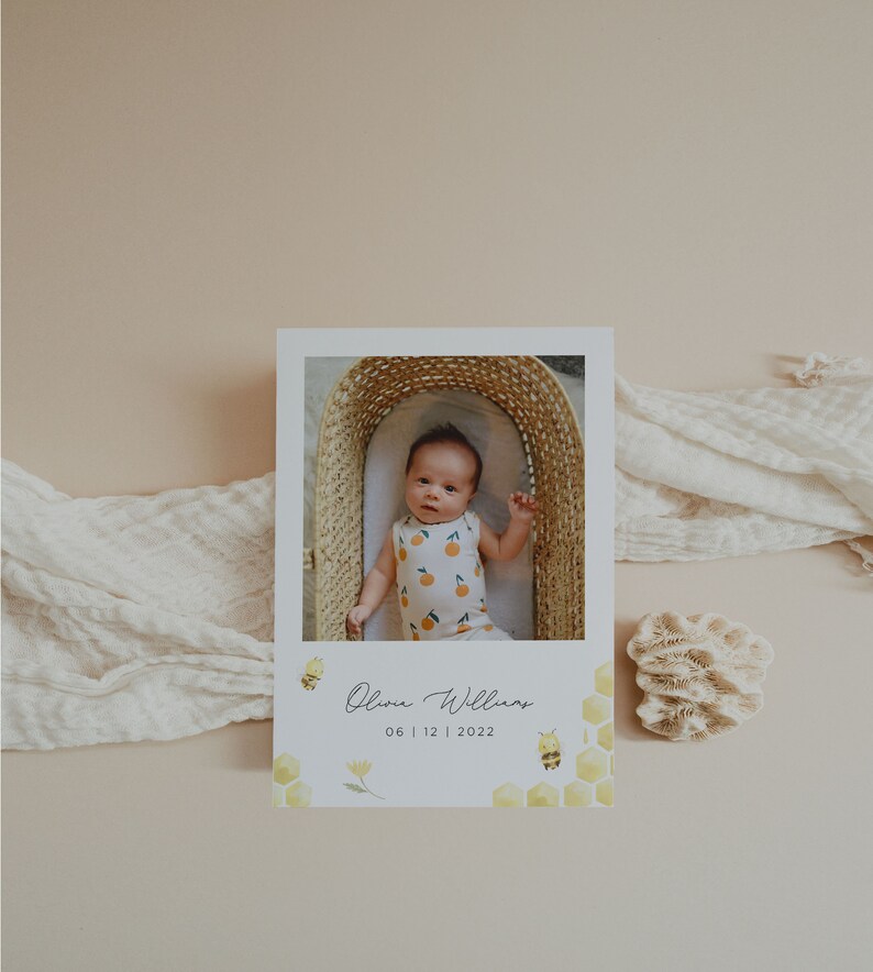 Op de afbeelding: Een witte kaart met een foto van een baby in een rieten mand. De kaart heeft een geel honingraatpatroon met bijen en bloemen. De tekst op de kaart luidt "Olivia Williams 06 | 12 | 2022".