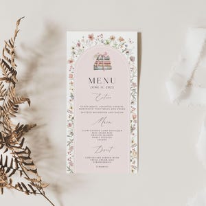 May include: A pink and white wedding menu with a floral border. The menu features a watercolor illustration of a stack of books. The text reads "MENU" and "JUNE 11, 2022".