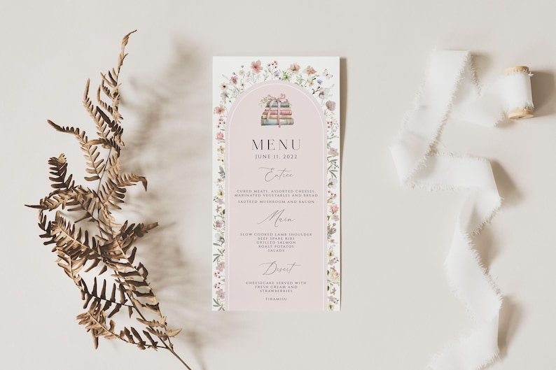 May include: A pink and white wedding menu with a floral border. The menu features a watercolor illustration of a stack of books. The text reads "MENU" and "JUNE 11, 2022".