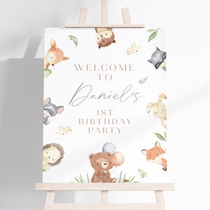 May include: A white welcome sign with a woodland theme. The sign features a variety of animals including a fox, a bear, a raccoon, a deer, a hedgehog, a rabbit, and an owl. The sign says "Welcome to Daniel's 1st Birthday Party".