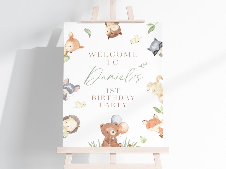 May include: A white welcome sign with a woodland theme. The sign features a variety of animals including a fox, a bear, a raccoon, a deer, a hedgehog, a rabbit, and an owl. The sign says "Welcome to Daniel's 1st Birthday Party".