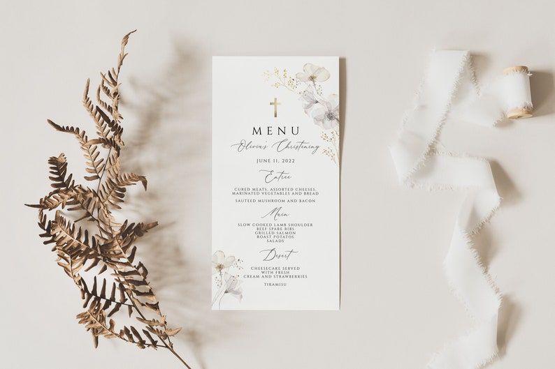 May include: A white menu card with a gold cross and floral design. The menu card lists the food served at Olive's Christening on June 11, 2023. The menu includes appetizers, main courses, and desserts.