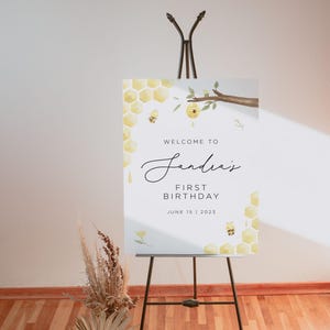 Może przedstawiać: Witamy na "Andreas' First Birthday" tablica z motywem plastra miodu i pszcz&oacute;ł. Tablica stoi na czarnym sztalugach i ma białe tło z ż&oacute;łtymi i brązowymi akcentami.