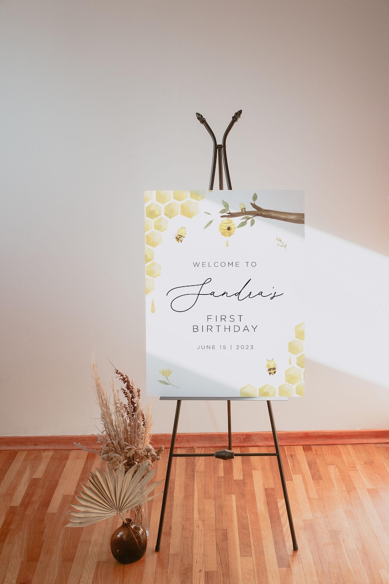Może przedstawiać: Witamy na "Andreas' First Birthday" tablica z motywem plastra miodu i pszcz&oacute;ł. Tablica stoi na czarnym sztalugach i ma białe tło z ż&oacute;łtymi i brązowymi akcentami.
