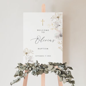May include: A white welcome sign with gold accents and floral designs. The sign reads "Welcome to Olivia's Baptism, September 17, 2022".