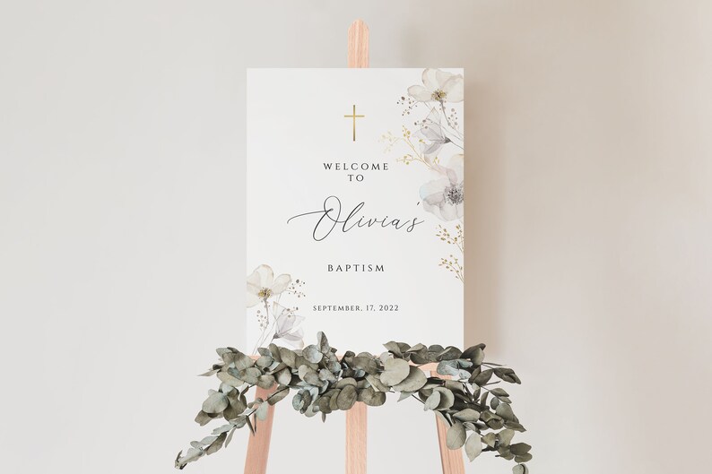 May include: A white welcome sign with gold accents and floral designs. The sign reads "Welcome to Olivia's Baptism, September 17, 2022".