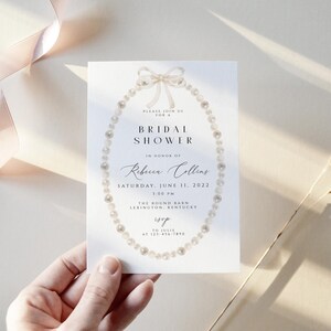 May include: A white bridal shower invitation featuring a pearl necklace and bow design. The invitation reads "Please join us for a Bridal Shower in honour of Rebecca Collins, Saturday, June 11, 2022, 5:00 PM, The Round Barn, Verington, Kentucky, RSVP at 123-456-7890".