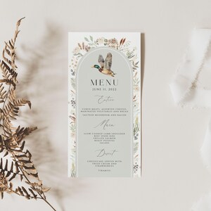 May include: A light gray wedding menu card with a watercolor illustration of a duck flying in an archway of floral greenery. The menu card has the text "MENU" and "JUNE 11, 2022" at the top. The menu includes the headings "Entr&eacute;e", "Main", and "Dessert".