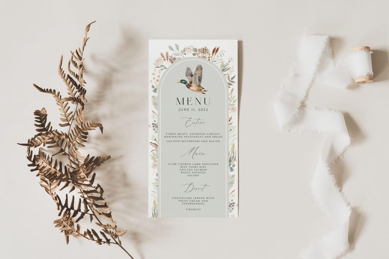 May include: A light gray wedding menu card with a watercolor illustration of a duck flying in an archway of floral greenery. The menu card has the text "MENU" and "JUNE 11, 2022" at the top. The menu includes the headings "Entr&eacute;e", "Main", and "Dessert".