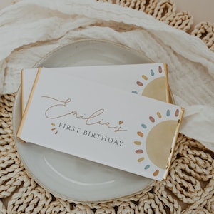 May include: A white card with gold trim and a colorful sun design. The card says "Emilia's First Birthday".