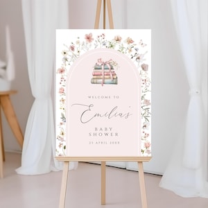 May include: A pink and white welcome sign with a floral border and a stack of books with a pink ribbon. The sign says "Welcome to Emilia's Baby Shower 25 April 20XX".