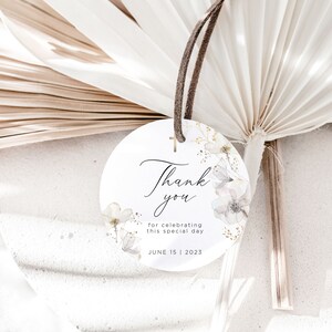 Puede incluir: Una etiqueta redonda y blanca con las palabras "Thank you" en escritura elegante, adornada con ilustraciones florales y una peque&ntilde;a cruz dorada. La etiqueta incluye el texto "for celebrating this special day" y la fecha "JUNE 15 | 2023". Una cuerda marr&oacute;n est&aacute; adjunta.