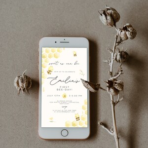 Op de afbeelding: Een smartphone die een gele en witte uitnodiging voor een eerste verjaardagspartij weergeeft. De uitnodiging heeft een honingraatpatroon en de tekst "sweet as can be" en "Emilia's First Bee-Day!" met een bij-illustratie. De uitnodiging bevat de datum, tijd en locatie van het feest.