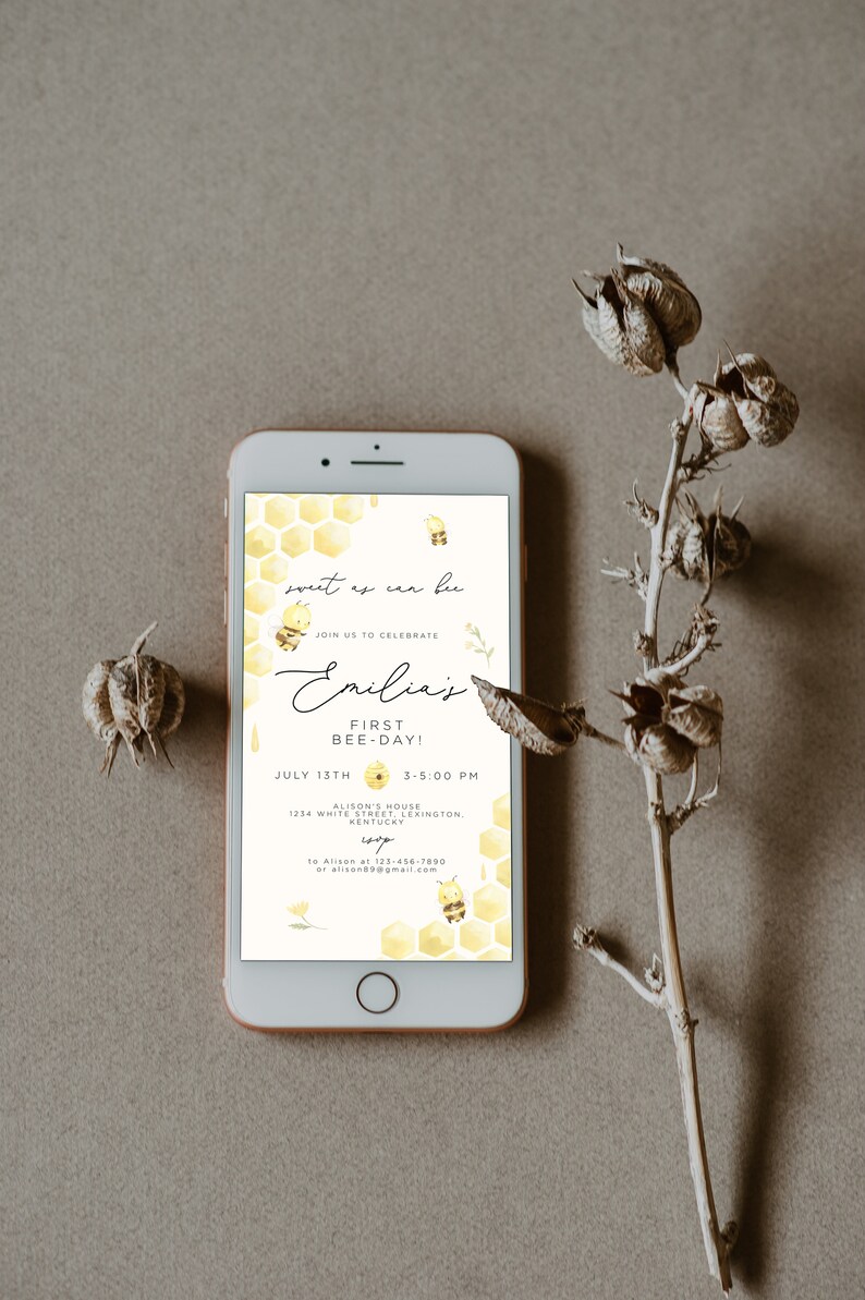 Op de afbeelding: Een smartphone die een gele en witte uitnodiging voor een eerste verjaardagspartij weergeeft. De uitnodiging heeft een honingraatpatroon en de tekst "sweet as can be" en "Emilia's First Bee-Day!" met een bij-illustratie. De uitnodiging bevat de datum, tijd en locatie van het feest.
