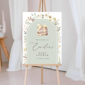 May include: A welcome sign for a baby shower with a green background and floral accents. The sign says "Welcome to Emilia's Baby Shower 25 April 20XX" and features a stack of books with a ribbon.
