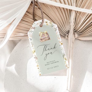 May include: A green and white thank you tag with a floral design. The tag features a stack of books and the text "Thank you" in cursive. The text "Olivia's Baby Shower 07/12/2023" is also included.