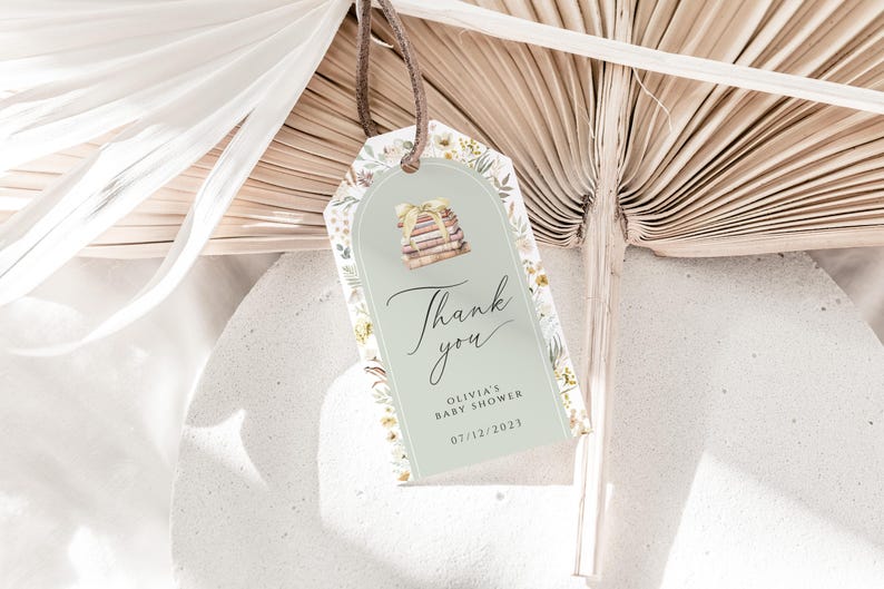 May include: A green and white thank you tag with a floral design. The tag features a stack of books and the text "Thank you" in cursive. The text "Olivia's Baby Shower 07/12/2023" is also included.