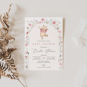 Pode incluir: Um convite para ch&aacute; de beb&ecirc; com Ursinho Pooh, rodeado por um arco floral. O convite inclui o texto "Please join us for a Baby Shower honoring Emilia Johnson" e detalhes do evento. Um ramo de samambaia seca e fita branca tamb&eacute;m est&atilde;o na imagem.